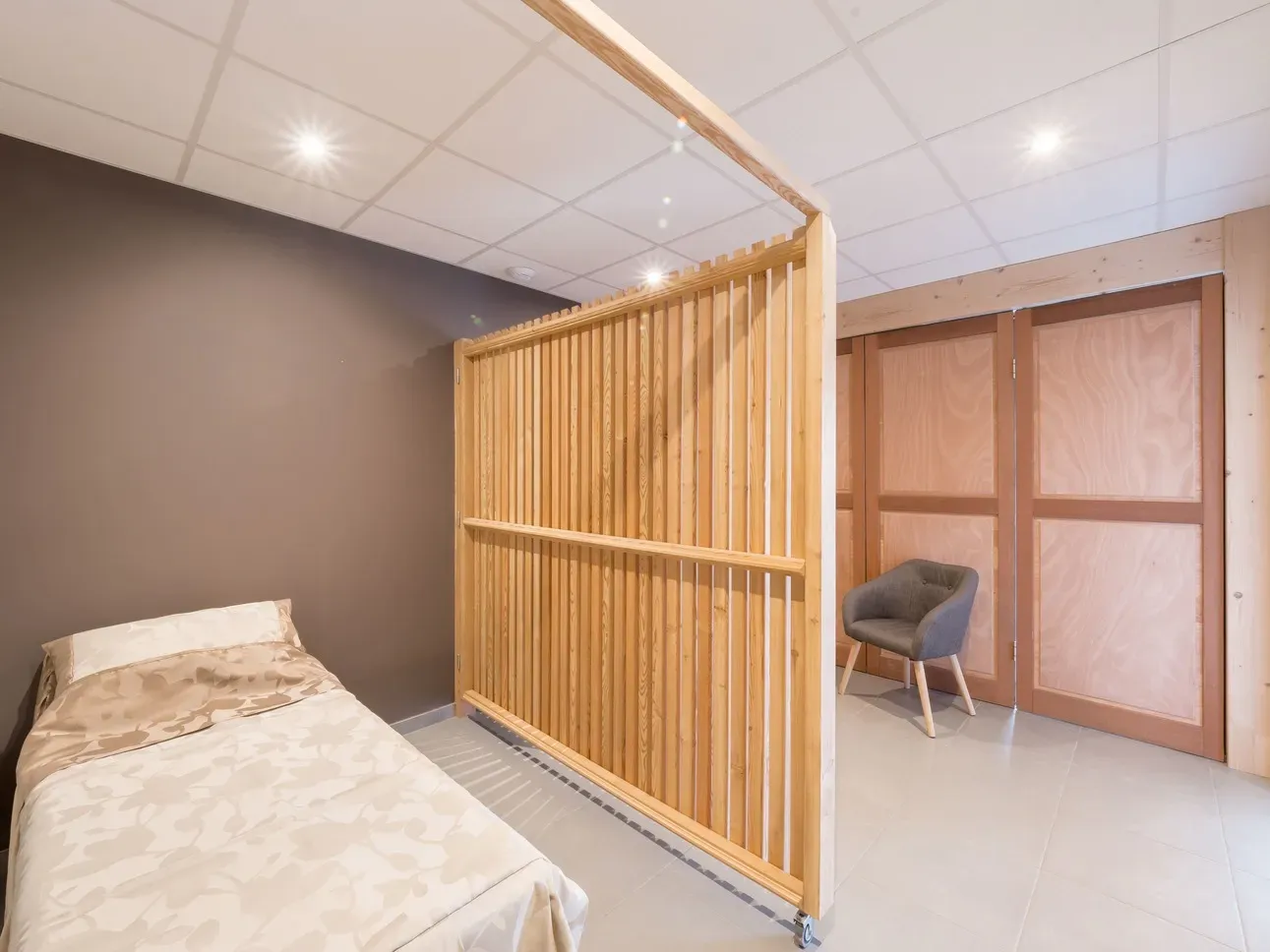 Chambre funéraire aménagée avec lit de repos, fauteuil et paravent en tasseaux de bois.